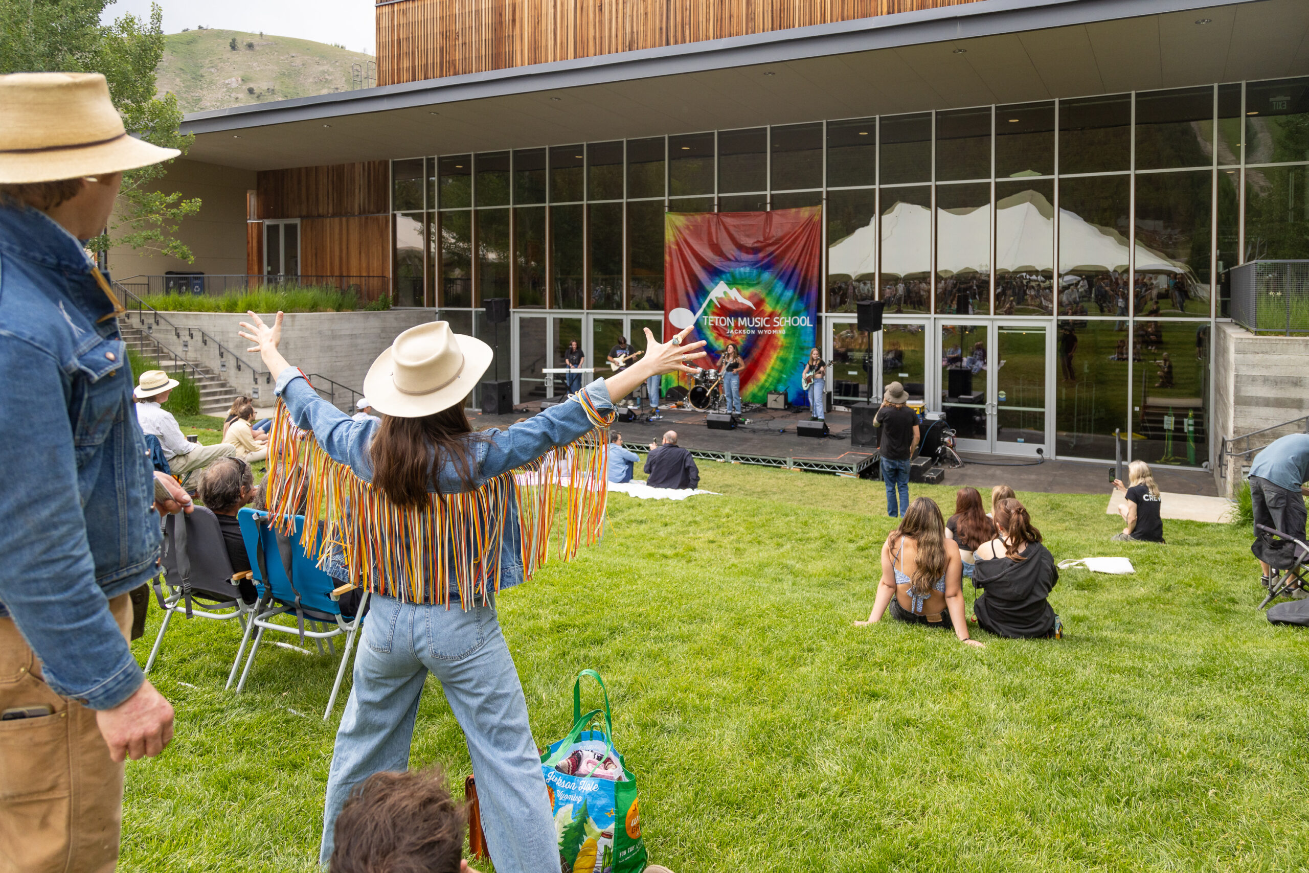 A person wearing a cowboy hat and denim jacket with frills with their arms stretched out wide watching a performance outside