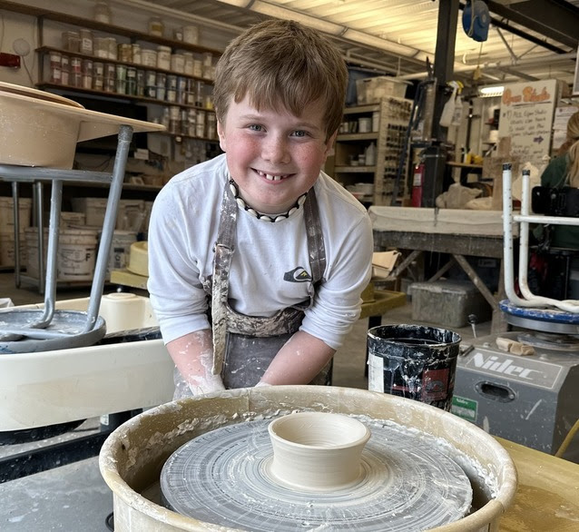 A child smiling stood in front of a pottery wheel