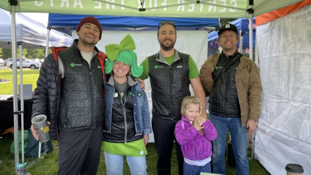 Three men, a woman and a small child under a tent, wearing Jackson Hole Center for the Arts down vests at Old Bill's Fun Run for donations. The woman is wearing a green foam wig.