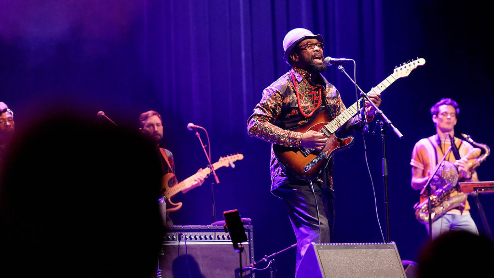 A man plays a brown electric guitar while singing on stage, he is wearing a bowler hat, glasses, and paisley pattern shirt with a large red bead necklace ,his band members are in the background playing a bass guitar and a saxophone