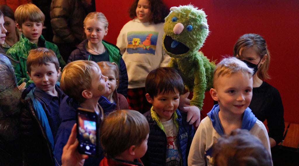 a group of small children of varying ages are gathering around a green fluffy puppet with blue eyes, a round pink nose, a blue tongue and pink hands.