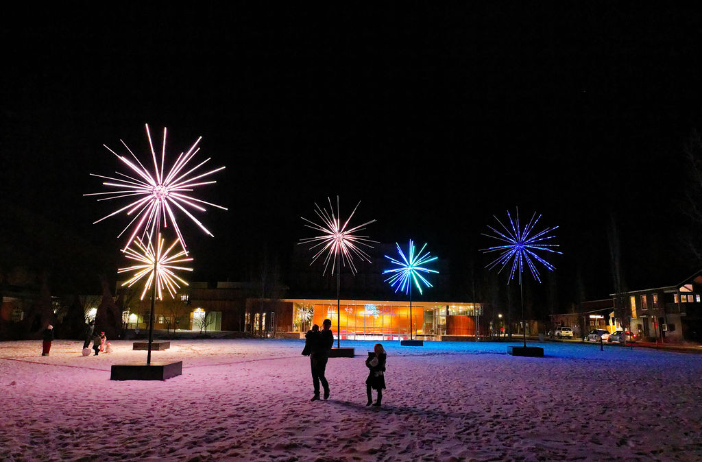 five large light sculptures in an open area with snow on the ground and two small families walking below them, they are brightly coloured and resemble fireworks or flowers