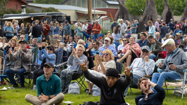 A crowd of people, mainly sat on garden chairs cheering while watching an outdoor performance. The two people at the front sat on the grass have their hands in the air