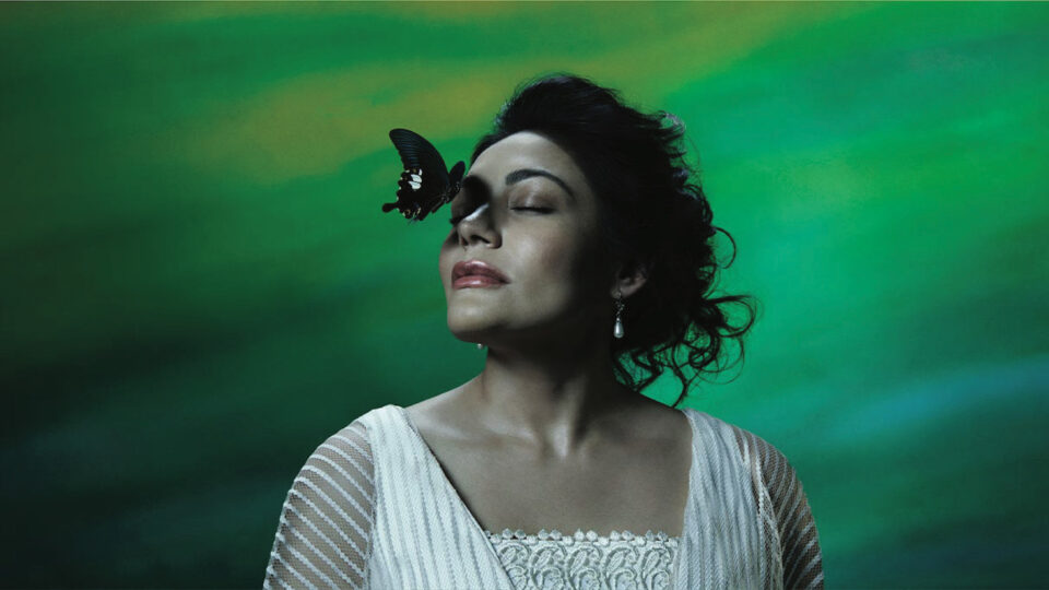 a beautiful woman wearing a white lace dress, teardrop pearl earrings and curly black hair pinned in an updo, with her eyes closed as a butterfly is about to land on her nose. she is in front of a green background.