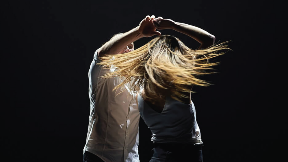 A couple dancing in front of a black background, a man spins a woman round as her long blonde hair swirls in different directions as she is moving