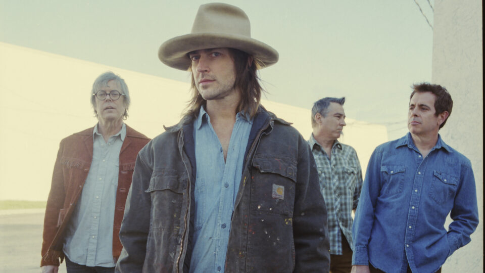 The band OLD 97's, a band of four middle aged male members wearing blue denim shirts, the front man is wearing a large beige hat and carhart jacket and has shoulder length shaggy brown hair