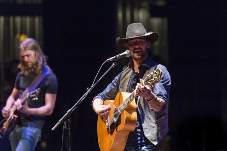 Two people performing on stage, one is out of focus. The person at the front is wearing a cowboy hat, playing the guitar and singing
