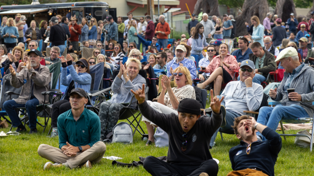 A crowd of people, mainly sat on garden chairs cheering while watching an outdoor performance. The two people at the front sat on the grass have their hands in the air